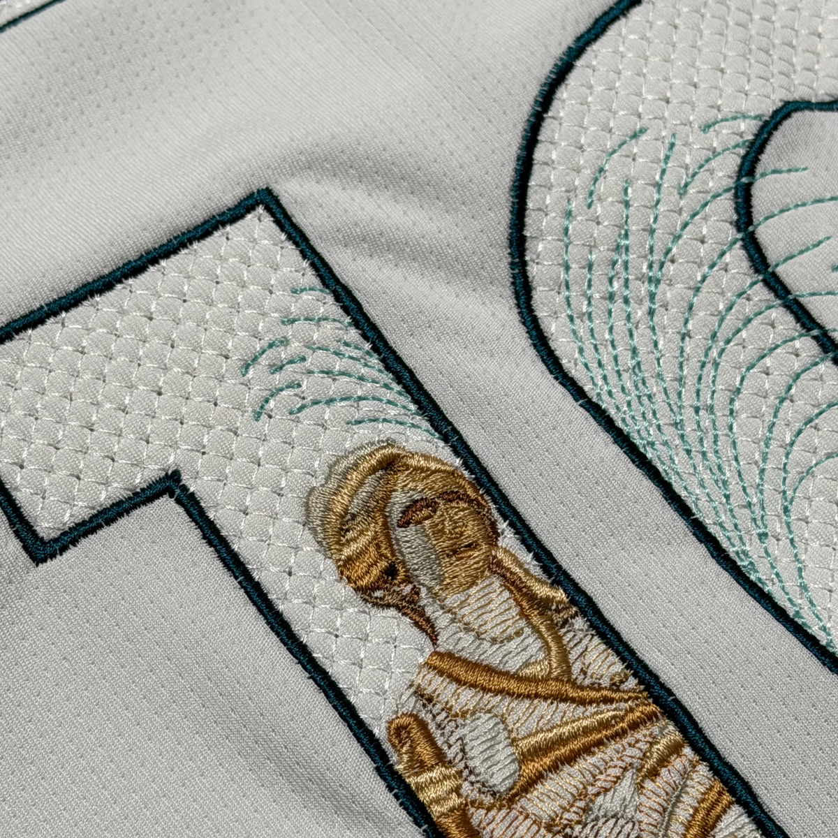 Macro detail of the Cibeles statue embroidered in gold thread, with the fountain spray stitched in green across the numbers