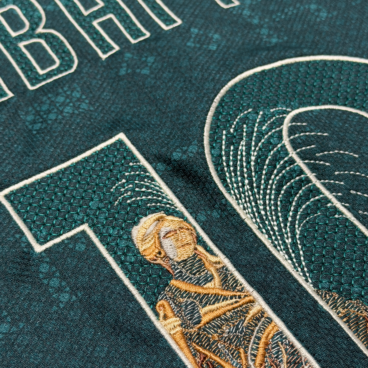 Macro detail of the Cibeles statue embroidered in gold thread, with the fountain spray stitched in cream across the numbers