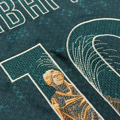 Macro detail of the Cibeles statue embroidered in gold thread, with the fountain spray stitched in cream across the numbers
