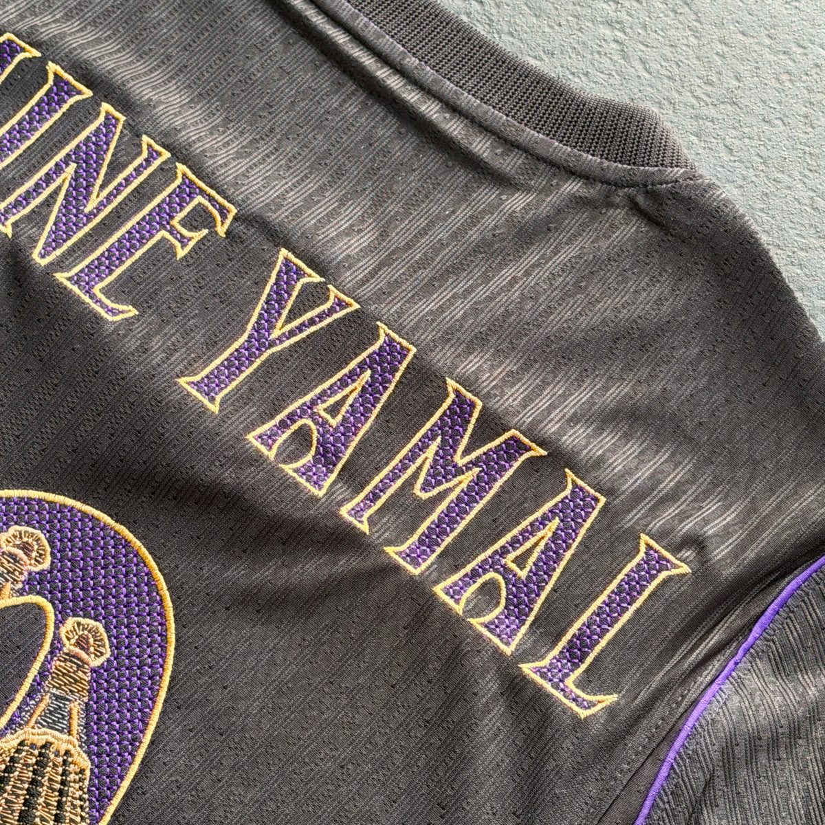 Detail of YAMAL in gold-outlined modernista lettering, stitched over the jersey's textured knit